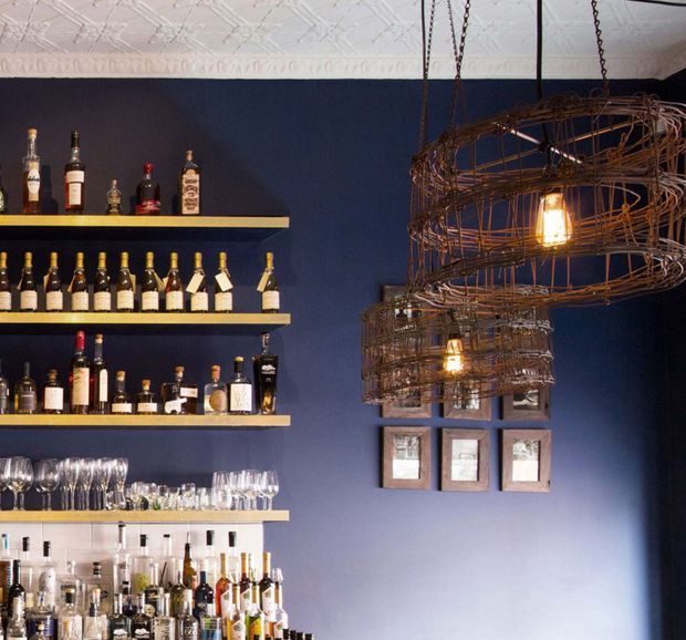 A dark, sophisticated home bar with deep navy walls, multiple open shelves displaying liquor bottles and glassware, and a light-coloured tiled or patterned ceiling. Large suspended sculptural wire-cage pendant lights with exposed bulbs dominate the right side, while framed artwork hangs below them.