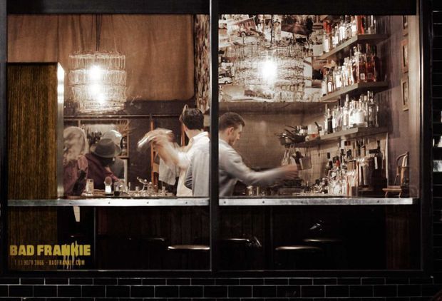 A dark, atmospheric bar interior viewed through large black-framed windows. The compact space features bartenders serving behind a bar, extensive illuminated bottle shelving, reflective mirrored surfaces, black subway-tile wall cladding, dark timber or wood-toned finishes, and a distinctive hanging glass pendant light. Text on the window identifies the venue as Bad Frankie.