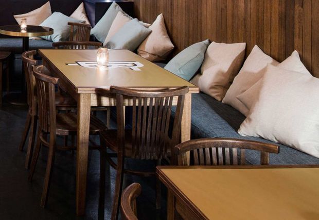 A warmly lit dining or hospitality seating area with long wooden tables, dark timber slat-back chairs, and built-in upholstered bench seating against a vertical timber-panelled wall. Neutral cushions and small table lamps create a cozy, refined atmosphere.