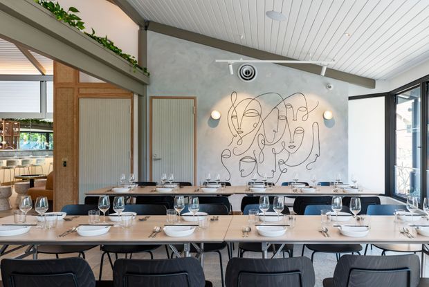 Contemporary restaurant dining room with long communal tables set with crockery and wine glasses. The space features a pale concrete-look feature wall with a large abstract line-art mural, timber-framed ribbed-glass doors, exposed dark structural beams, a white slatted ceiling, pendant and wall lighting, and large glazed openings to the side.