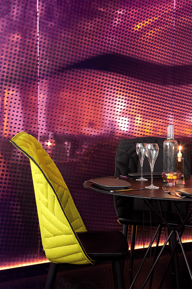 A dramatic, highly stylised dining nook with a round dark dining table, black upholstered dining chairs, and a vivid chartreuse-yellow sculptural chair. The walls and ceiling are covered in a continuous purple-magenta dotted feature finish with reflective, graphic patterning. Glassware, candles, and warm concealed lighting create an intimate, glamorous atmosphere.