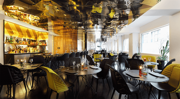 A stylish contemporary bar and dining venue with a long open dining area, extensive liquor shelving and a dark bar counter along the left wall. Numerous small round tables are arranged with upholstered dining chairs, including mustard-yellow accent chairs and dark seating. The space features a highly reflective black mirrored ceiling, warm timber wall cladding, geometric patterned wall panels, large windows on the right, and dramatic pendant lighting. The overall atmosphere is sophisticated, lively, and luxurious.