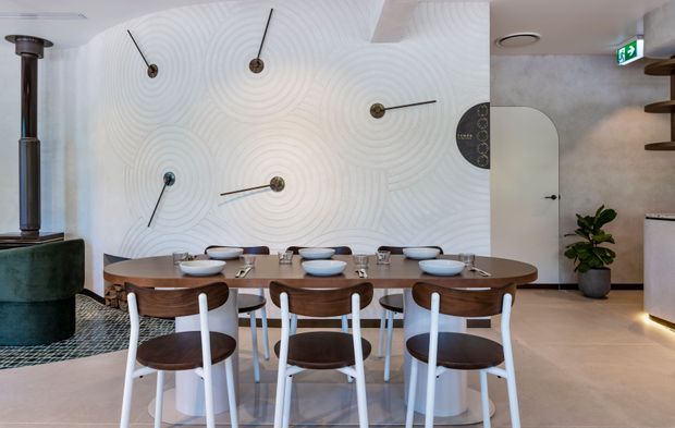 Contemporary dining area with a long rounded-edge timber dining table and sculptural mixed-material chairs, set against a white textured feature wall with concentric relief patterns and minimalist wall-mounted lighting. The room includes a curved white doorway, pale stone or tiled flooring, a dark green cylindrical column or fireplace element, a small indoor plant, and partial built-in shelving. The overall scheme is refined, sculptural, and neutral with warm timber accents.