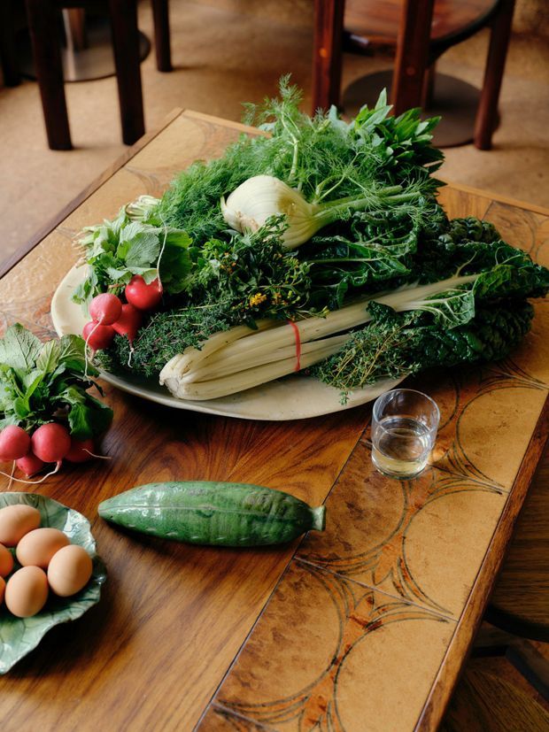 A close-up still-life of freshly harvested vegetables arranged on a large ceramic platter atop a richly finished wooden dining table. Visible produce includes fennel, radishes, leafy greens, celery, herbs, cucumber, and eggs, with a drinking glass nearby. Dark timber dining chairs and a warm, patterned tile or inlaid tabletop border are visible in the background. The image is food-focused rather than a clear architectural space.