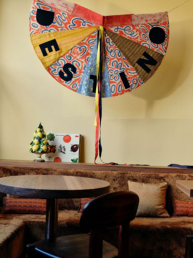 A warmly lit interior dining and lounge area with a round timber table and dark chairs in the foreground. A built-in upholstered banquette runs along the back wall with patterned cushions, while a colourful handmade textile wall hanging and small decorative objects sit above it. The room has a cozy, eclectic, craft-focused character with earthy tones and playful primary-colour accents.
