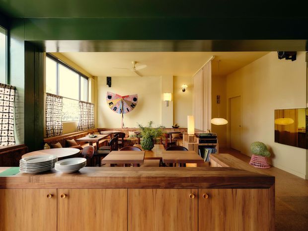 Warmly lit open-plan dining and living space viewed across a large timber kitchen or serving island. The room features extensive glazing with patterned roller blinds, timber dining tables and chairs, built-in shelving, pendant and wall lighting, indoor plants, and a colourful wall artwork. A dark green structural beam or ceiling surround contrasts with the warm timber cabinetry and cream walls.