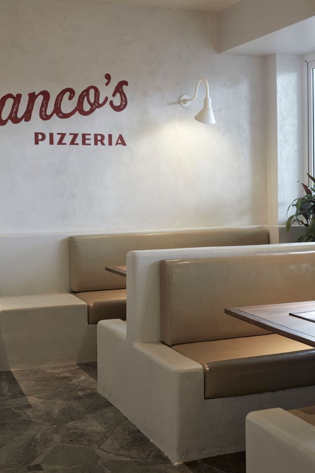 Interior view of a pizzeria dining area with built-in upholstered booth seating, warm beige and cream finishes, a textured plaster wall with red “Pizzeria” lettering, a white wall-mounted sconce, dark irregular stone flooring, and a window with indoor planting.