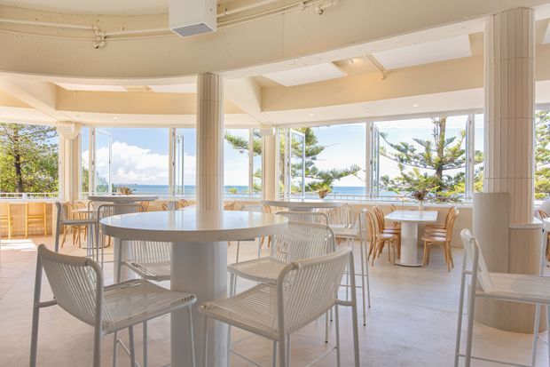 Bright, airy coastal café or restaurant interior with extensive floor-to-ceiling glazing overlooking the ocean. The open dining area features round white tables, light woven chairs, pale tiled flooring, fluted cylindrical columns, exposed ceiling services, and abundant natural light.