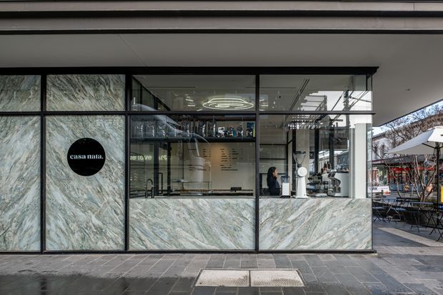 Street-facing commercial café frontage with extensive black-framed glazing and dramatic veined stone panels. The interior shows a service counter, display shelving, pendant lighting, and café equipment, while outdoor seating is visible to the right. The design has a refined contemporary character with a dark, graphic palette and natural stone feature surfaces.