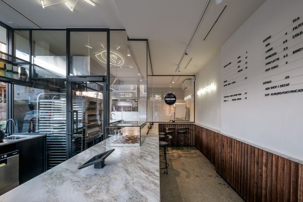 Contemporary commercial food-service interior with a long pale stone counter, glass-fronted kitchen and display areas, dark-framed glazing, timber wall panelling, stools, and a white menu wall. Linear recessed lighting and clean modern detailing create an open, refined café or takeaway environment.