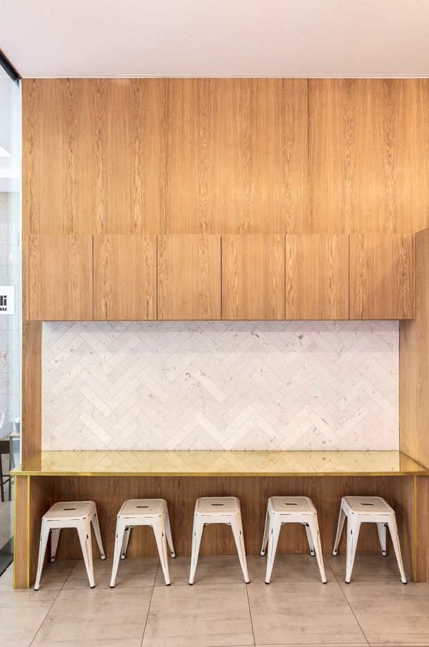 A contemporary built-in dining or casual seating nook featuring warm timber-look cabinetry and wall panelling, a long integrated bench, and five white metal stools. The backsplash is clad in a pale herringbone-patterned tile, with a tiled floor and a simple pale ceiling. The composition appears to be in a commercial or hospitality setting, viewed straight on.