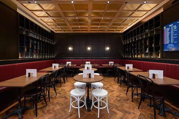 A stylish restaurant dining room with rows of timber tables and black bentwood-style chairs arranged around a central communal table. Deep red upholstered banquette seating lines the walls, while black open shelving displays bottles and objects. The room features a warm parquet timber floor, dark wall panelling, and a prominent coffered timber ceiling with recessed spotlights. A digital menu screen is visible on the right wall.