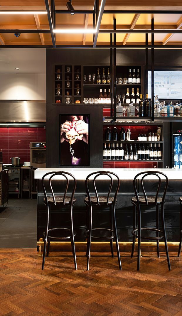A sophisticated bar and hospitality interior with a long dark bar counter, three tall curved-back bar stools, extensive open bottle shelving, and a back-of-house kitchen area. The space combines dark timber or black cabinetry, a glossy red tiled splashback, stainless-steel kitchen surfaces, warm timber ceiling panels, dramatic integrated lighting, and a richly patterned timber parquet floor. A framed digital artwork and drinks refrigeration reinforce the commercial bar setting.