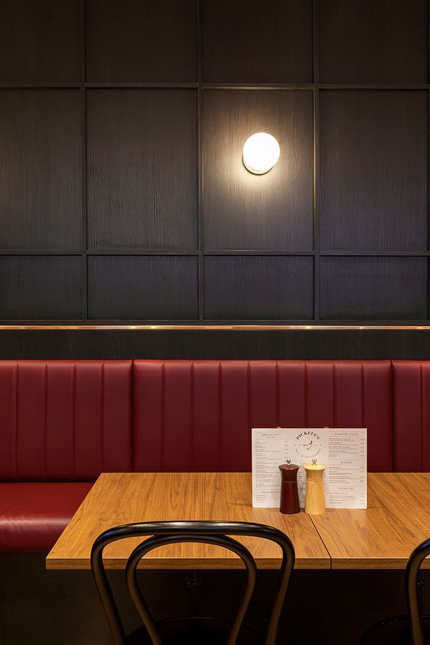 A moody restaurant dining area with a long red upholstered banquette, warm timber tables, dark vertically textured wall panelling, black bentwood-style chairs, and a round wall light. Menus and table condiments sit on the table, clearly indicating a hospitality setting.