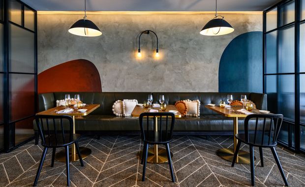 A stylish restaurant or hospitality dining area with a long black banquette, several set dining tables, black timber chairs, dramatic pendant lights, and a textured concrete-look feature wall. Large abstract arch-shaped wall colour blocks in rust orange and deep blue add bold contrast, while black-framed fluted or ribbed glass partitions enclose the sides. The space has a moody, refined, contemporary feel.