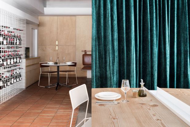 A stylish dining area with a long light timber dining table set with white plates, cutlery, wine glasses and a small vase. Teal velvet curtains form a dramatic backdrop, while the adjoining space features terracotta tiled flooring, light timber wall cabinetry, a compact round dining table, leather seating and a white mesh screen or partition. The interior combines warm natural materials with bold jewel-toned textiles and contemporary detailing.