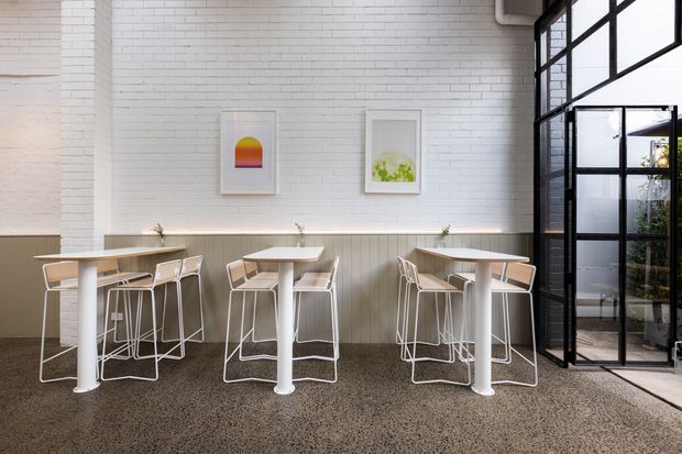 A bright contemporary café or commercial dining space with white painted brick walls, vertical wall panelling, terrazzo flooring, and a black-framed glazed partition. Several small white tables are paired with light wood and white bar stools, while minimalist framed artwork adds colour to the walls.