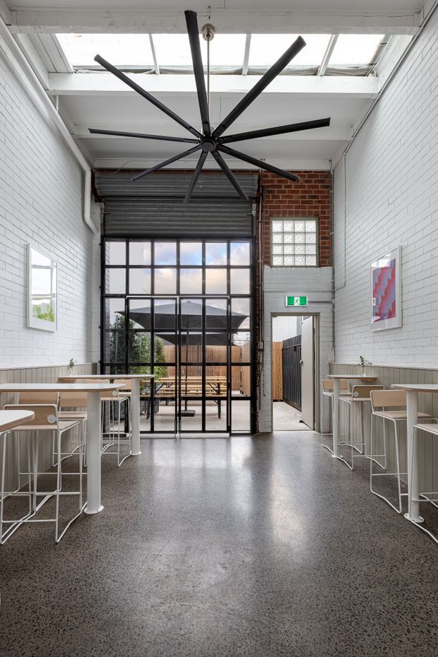 A bright, narrow commercial-style interior with white-painted brick walls, polished exposed aggregate or speckled concrete flooring, and café-style high tables and stools arranged along both sides. A large black industrial ceiling fan hangs beneath a glazed skylight roof, while black-framed industrial glazing and a roller door open toward an outdoor seating area. A red brick accent wall, glass-block window, artwork, and an exit door add character to the space.