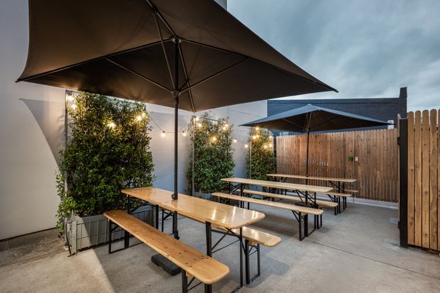 A covered outdoor courtyard dining area with long timber picnic tables and benches, large dark umbrellas, potted screening greenery, festoon lights, concrete paving, and a timber privacy fence. The space appears suited to casual communal dining or entertaining.