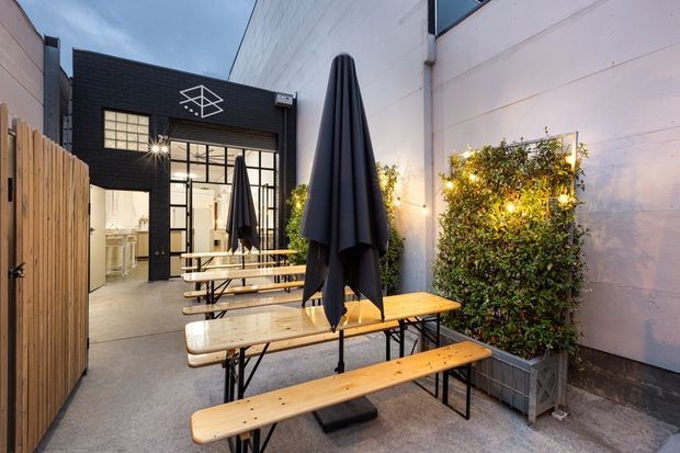 A narrow urban courtyard or laneway dining area with timber picnic benches, closed black market umbrellas, festoon lighting, planter boxes, and a timber privacy fence. The space opens into a modern black-brick commercial interior with large industrial-style glazing and a visible kitchen or service area. The setting is photographed at dusk with warm lighting and a casual hospitality atmosphere.