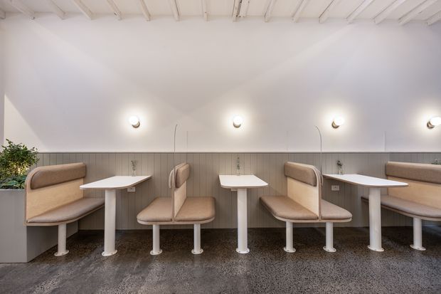 A bright, modern commercial dining or café interior with a long built-in banquette, multiple upholstered booth seats, white pedestal tables, vertical wall panelling, exposed white ceiling beams, and wall-mounted globe lights. The space has a clean, minimal fit-out with a polished concrete or terrazzo-like dark floor and abundant natural light.