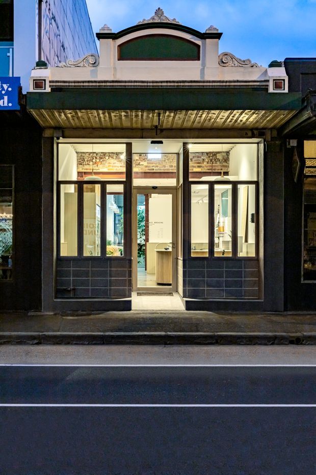Street-facing view of a narrow, renovated commercial storefront in a heritage-style building. The facade combines a dark shopfront with glazed entry doors, tiled lower panels, a projecting awning, and an ornate cream architectural parapet with decorative scrollwork and a curved central panel. Warm interior lighting reveals a compact retail or service space with shelving and a counter behind the glazing.