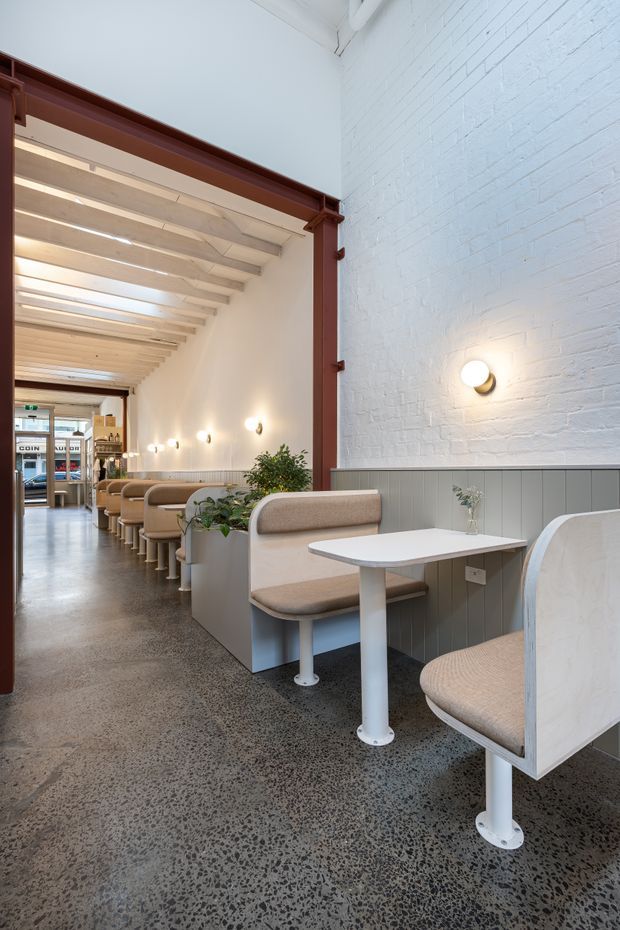A bright contemporary café interior in a long, narrow space. Repeated upholstered booth seating and small white pedestal tables line the white-painted brick wall, with pale grey-green wainscoting, warm wall sconces, polished terrazzo flooring, and exposed white-painted ceiling beams. A large dark red structural frame defines the opening to the seating area.