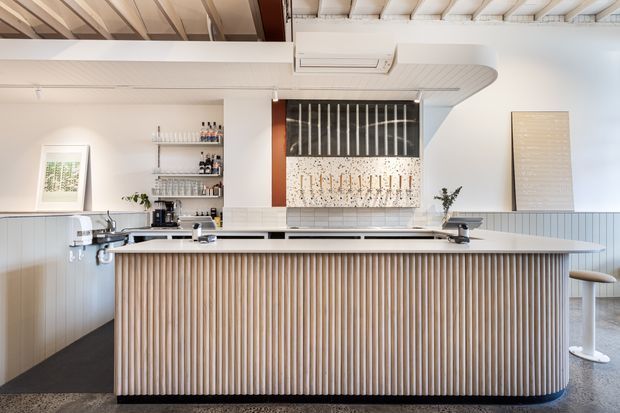 Bright contemporary kitchen with a large island featuring a vertically slatted timber-clad base and pale stone benchtop. White cabinetry and walls are paired with open shelving, integrated appliances, a ribbed ceiling, and a dark patterned feature splashback. The space has a clean, refined hospitality-inspired feel.