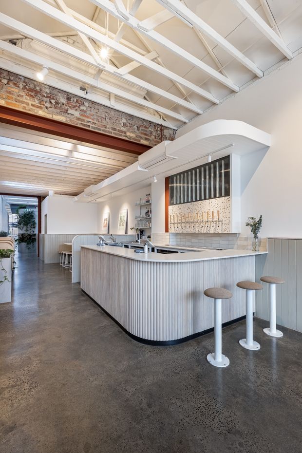 Bright commercial hospitality interior featuring a curved ribbed service counter with white stone benchtops, stools, and integrated display shelving. The space combines exposed painted roof trusses, a reclaimed exposed-brick wall, timber-lined ceiling sections, polished concrete flooring, and a distinctive tiled and ribbed feature wall behind the counter.