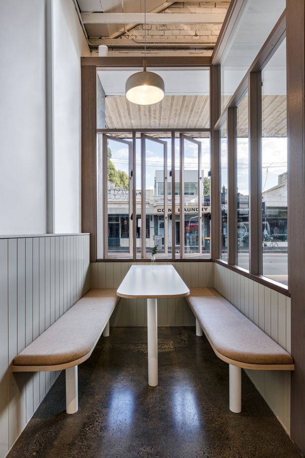A compact cafe dining nook with built-in upholstered bench seating along two walls, a small white table, full-height timber-framed windows, and views to a commercial street outside. The space combines pale vertical wall panelling with a polished dark concrete floor, exposed painted ceiling structure and masonry, and a suspended dome pendant light.