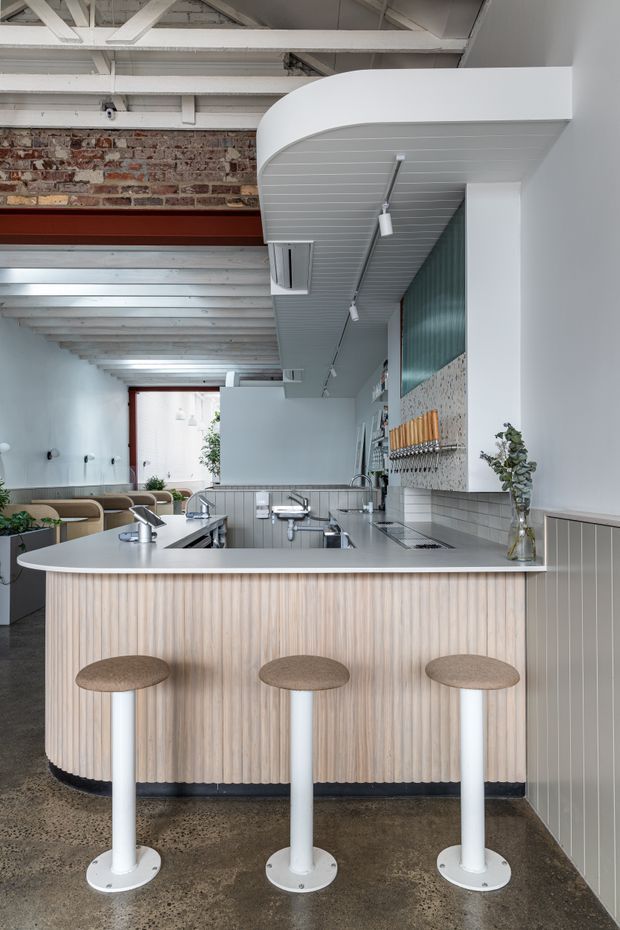 A bright contemporary café or hospitality space centred on a curved island counter with a pale fluted timber front, light benchtop and three white pedestal stools. The room combines exposed white-painted structural beams and ceiling boards with an exposed brick wall, ribbed wall panelling, tiled backsplash, integrated cooking or service equipment, and track lighting. Additional seating is visible to the left, creating an airy, refined but industrial-inspired interior.