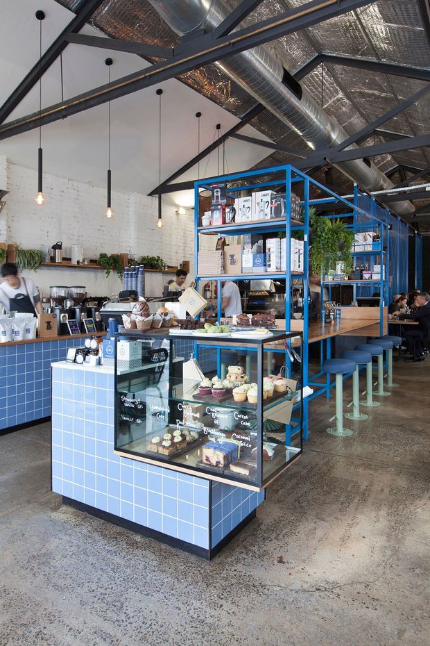 Interior photograph of a contemporary café with an industrial fit-out. The space features a blue tiled service counter and display island, blue steel shelving, timber counters, bar seating, exposed dark structural beams, visible silver HVAC ductwork, reflective insulated ceiling panels, white-painted brick walls, pendant lighting, and a polished concrete floor. Pastries, coffee equipment, shelving, and café seating are visible throughout.