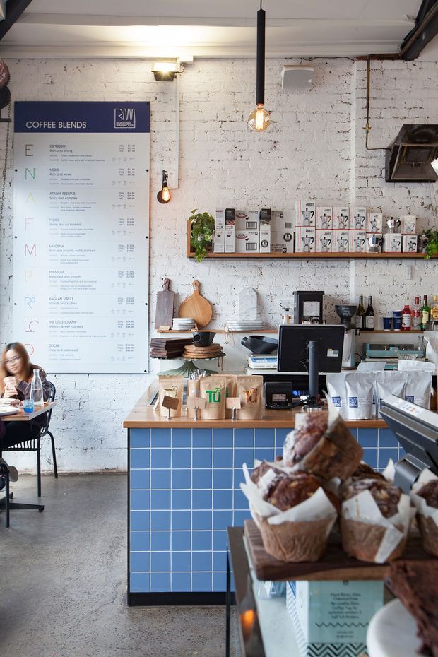 Interior photograph of a compact, industrial-style coffee shop with a white-painted exposed brick wall, visible coffee menu, service counter, espresso equipment, retail coffee products, baked goods display, timber shelving, and pendant lighting. The counter front is finished with blue square tiles, and the polished concrete floor and exposed ceiling services reinforce the café fit-out. A seated customer is partially visible at the left.