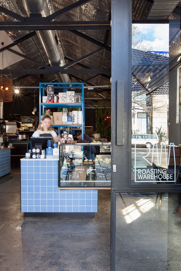 A compact industrial-style coffee shop viewed from the entrance, with a blue tiled service counter, glass display case, coffee equipment, and blue metal shelving. The space has exposed dark steel roof trusses, reflective roof insulation, visible ductwork, polished concrete flooring, and black-framed glazed doors. Strong daylight enters through the storefront, with café branding visible on the glass.