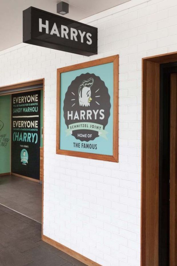 Interior view of a branded restaurant or food venue entry area. A white-painted brick wall features a black projecting sign reading “HARRYS,” a framed vintage-style “Harrys Schnitzel Joint” poster, and additional branded graphics visible through an opening. Dark tiled flooring, timber door and poster frames, and simple ceiling-mounted lighting complete the commercial fit-out.