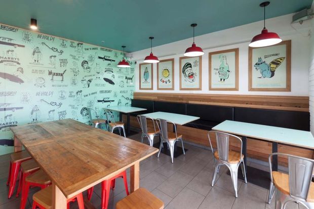 A casual café or dining space with a long communal timber table, smaller pale tables, metal chairs, and a built-in black banquette. The room features turquoise walls and ceiling, red pendant lights, a timber dado rail, and illustrated graphic wallpaper and framed artwork, creating a playful retro-industrial atmosphere.