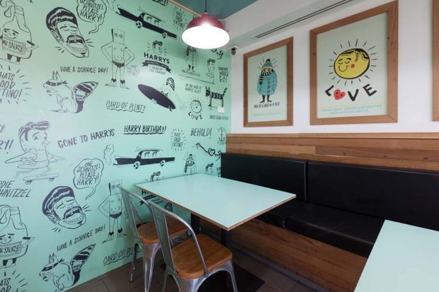 A compact retro-styled café or casual dining nook with a turquoise illustrated feature wall covered in hand-drawn cartoon graphics and lettering. A wall-mounted black upholstered banquette runs along a timber-clad wall beneath framed whimsical prints, paired with a pale turquoise table and metal dining chairs. A red pendant light hangs overhead, creating a playful, graphic hospitality interior.