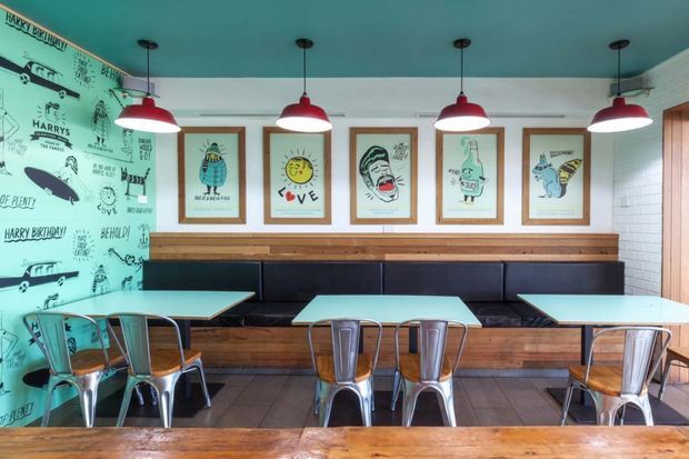 A brightly styled casual dining area with turquoise walls and ceiling, a long built-in black banquette, timber wall ledge and base, framed graphic artwork, and multiple red pendant lights. Small tables are paired with industrial-style metal chairs, creating a playful café or restaurant interior.