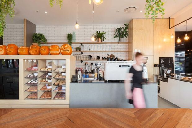 A bright, contemporary café interior viewed from the customer area, with a service counter, commercial coffee machine, pastry display cabinet, menu board, tiled backsplash, timber cabinetry, indoor plants, and warm pendant lighting. A herringbone timber floor and orange pumpkin display add colour and warmth.
