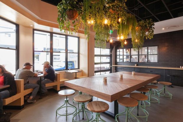 A bright, contemporary café or casual commercial dining space with a large communal timber table surrounded by round stools, booth seating along expansive windows, and a long black tiled counter at the rear. Hanging greenery and warm pendant lights decorate the exposed dark ceiling, while large glazed openings provide strong natural light.