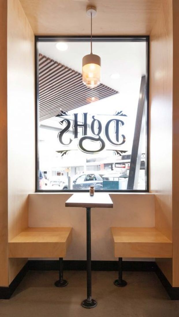 A compact commercial café seating nook viewed from inside, with two built-in light timber bench seats facing a narrow pedestal table beneath a pendant light. A large black-framed window overlooks the street and carries reversed café lettering, while the surrounding walls and ceiling are clad in warm-toned timber.