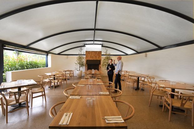 A spacious restaurant dining area with long and round timber tables, woven timber chairs, place settings, and a central fireplace clad in warm wood. The room has a curved translucent ceiling supported by dark exposed framing, pale plaster walls, polished concrete or terrazzo-like flooring, and extensive open glazing to greenery on one side. The atmosphere is warm, airy, and contemporary with natural materials.