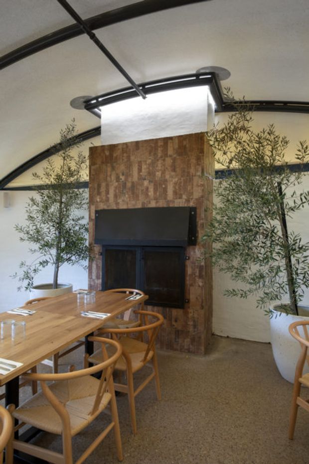 Interior view of a contemporary dining space with a long timber dining table and curved timber chairs. A central fireplace or dark built-in feature is set into a vertical reclaimed timber-clad wall, with large potted olive trees and curved black structural ceiling elements creating an organic, softly industrial atmosphere.