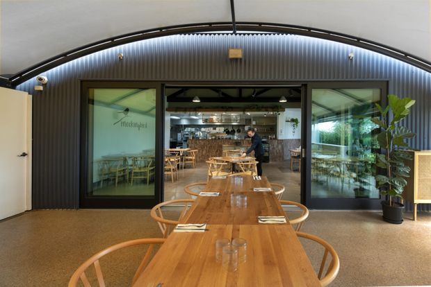 Interior view of a contemporary restaurant or café dining area, centred on a long solid timber communal table with curved wooden chairs. The space has a high arched ceiling, dark corrugated wall cladding, exposed dark structural framing, large glazed sliding openings, and a commercial kitchen and bar visible beyond. Potted greenery and warm pendant lighting add a welcoming hospitality atmosphere.