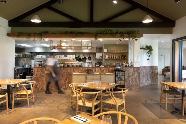 A rustic-modern café or restaurant interior with a prominent open commercial kitchen and service counter. The space features exposed dark timber roof trusses, white walls, polished concrete flooring, timber-framed dining tables and chairs, open timber shelving, hanging pendant lights, and greenery integrated along the upper shelving. A blurred staff member is moving behind the counter, while a glazed opening on the right reveals an adjoining seating area.