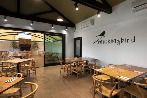 A warmly lit restaurant dining room with timber tables and curved-back wooden chairs arranged throughout the space. The room has polished concrete flooring, white walls, exposed dark structural beams, black track lighting, wall-mounted air-conditioning units, and large black-framed glazed openings connecting to an additional dining area. A prominent “Mockingbird” bird logo is displayed on the wall.