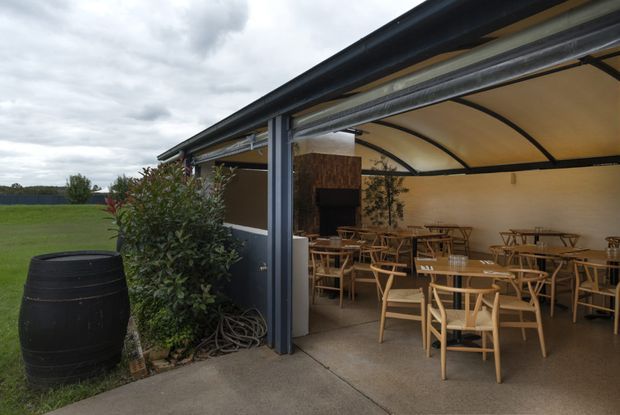 A covered outdoor dining and seating area beside an open grassy landscape. The structure has a dark metal frame, curved translucent canopy roof, concrete flooring, timber tables and chairs, and a dark outdoor fireplace or feature wall at the rear. Planting and a barrel add a rustic hospitality feel.