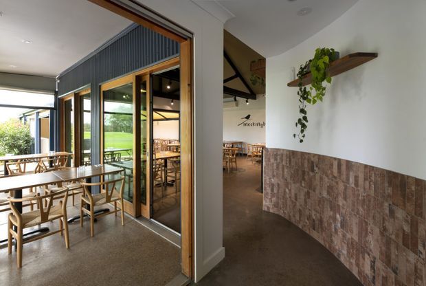 Interior view of a contemporary café or restaurant, showing a dining area with timber tables and chairs, full-height glazed folding or sliding doors opening toward an outdoor seating area, and a corridor-like entry zone. The space features polished concrete flooring, white plaster walls, timber-framed glazing, dark corrugated wall cladding, exposed dark structural beams, and a lower wall section finished in textured brown tile or stone. Floating timber shelves with trailing plants add a natural accent.