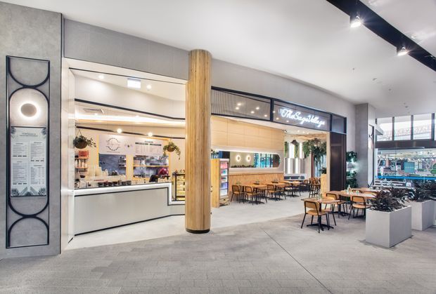 A bright contemporary commercial food and dining venue in an indoor retail or hospitality setting. The open storefront includes a service counter on the left, a central dining area with timber tables and chairs, overhead signage, dark-framed glazed partitions, a cylindrical timber-clad column, grey wall finishes, and polished track lighting. Additional retail or food-service space is visible to the right.