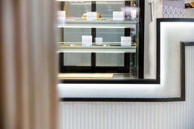 Close-up of a refined commercial display or retail interior featuring illuminated glass display shelving with small product labels, framed by dark trim and a textured pale wall panel. The foreground includes a softly blurred vertical architectural element, while the lower wall has vertical fluted detailing.